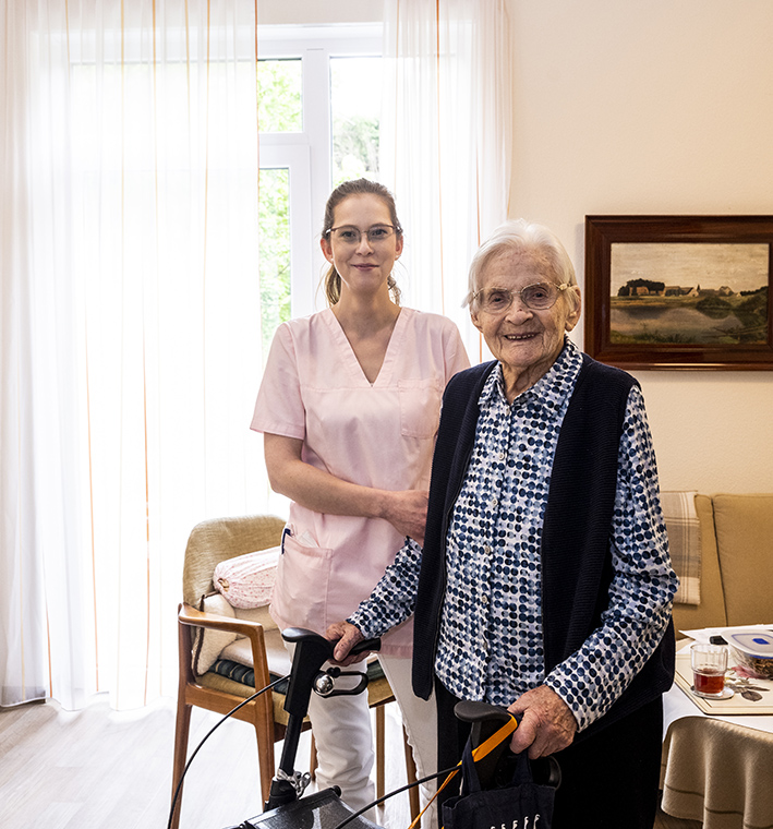 Pflegekraft steht mit Bewohnerin im Pflegeheim-Zimmer