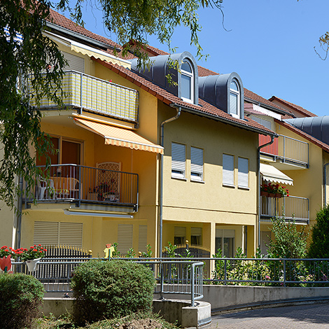 Standort avendi mobil Kehl, gelbe Häuser mit Balkonen, Grünflächen mit Sträuchern, Bäume spenden Schatten.