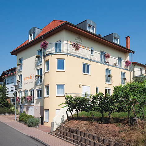 Standort Haus Burgblick von außen, Eingang zu mehrstöckigem gelbem Haus neben Weinbergen, oben ein großer Balkon.