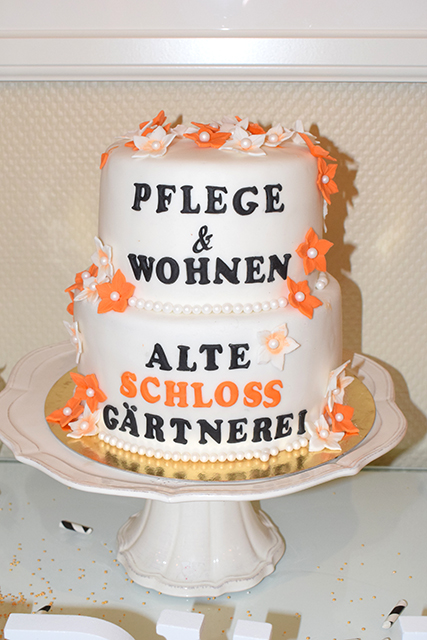 Zweischichtige Torte mit weißem Zuckerguss und orangenen Deko-Blüten, Aufschrift "Pflege & Wohnen alte Schlossgärtnerei"