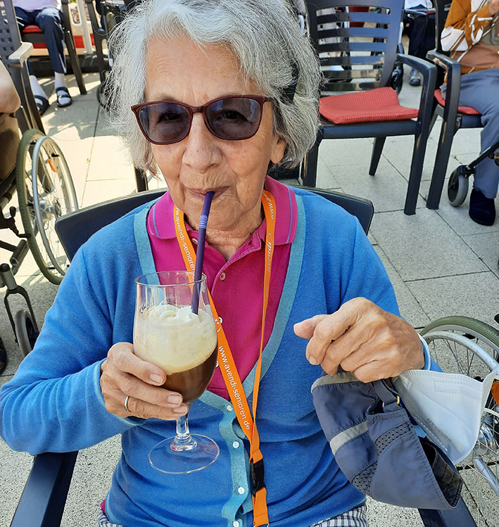 Eine Bewohnerin mit Sonnenbrille sitzt im Rollstuhl und genießt einen Eiskaffee mit Strohhalm.